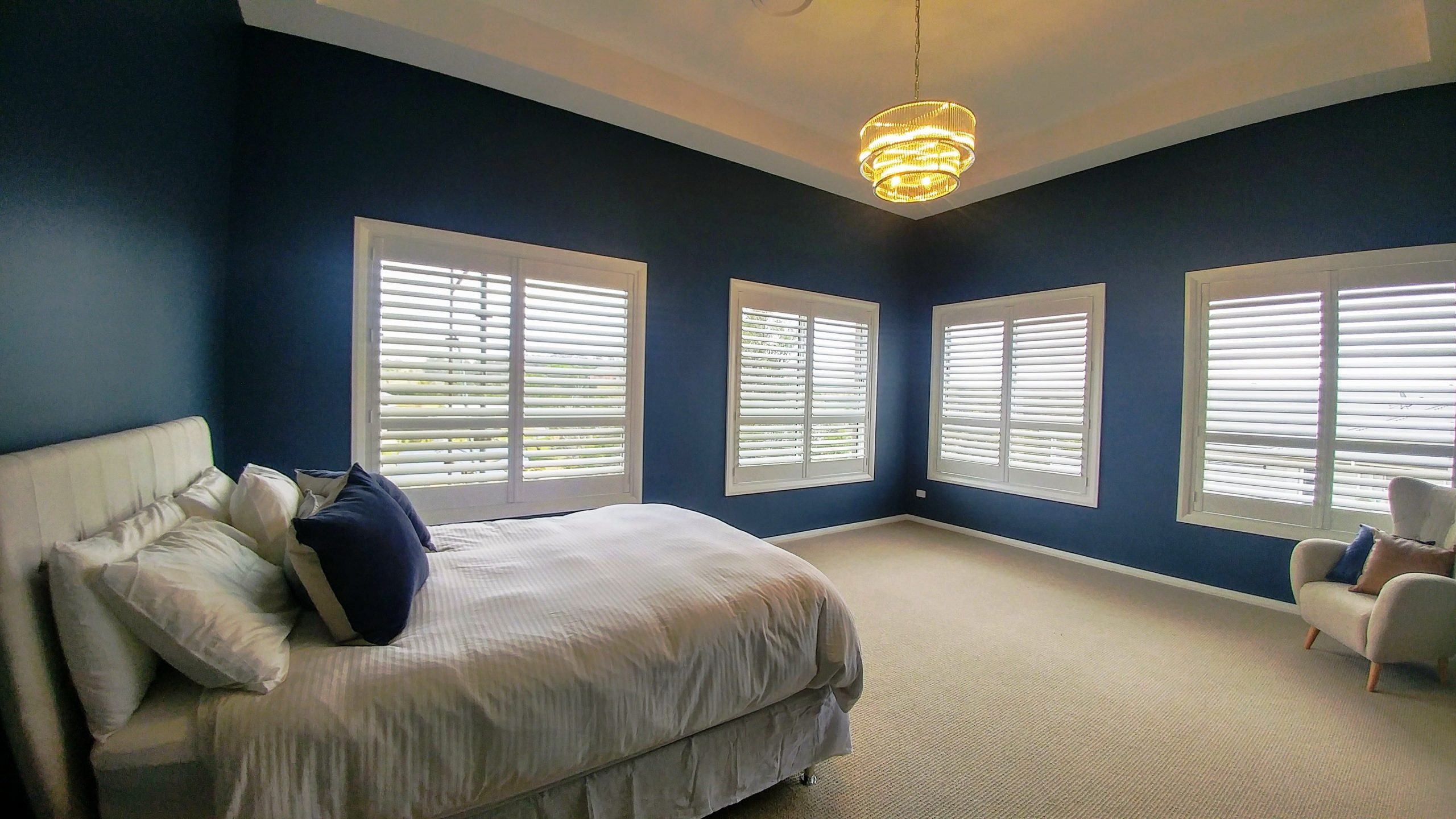 Spacious bedroom featuring stylish polymer shutters on windows, a plush bed with decorative pillows, and modern lighting against a deep blue wall.